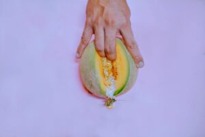 A hand gently touches a ripe melon on a pastel pink backdrop, creating a vivid and conceptual image.