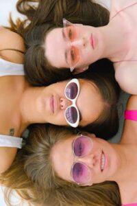 Three women wearing stylish sunglasses lie closely, showcasing friendship and fashion.