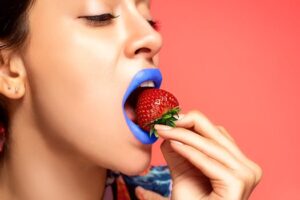 Colorful close-up of a woman with blue lipstick eating a ripe strawberry.